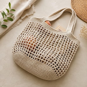 May include: A beige crochet tote bag featuring a solid base and a wide, open-grid mesh pattern throughout the main body. The bag includes two integrated handles and contains a small book and a round orange fruit visible through the mesh.