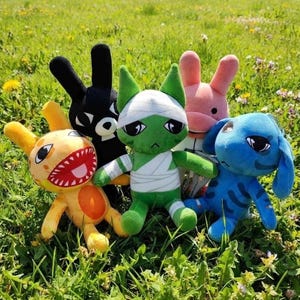 May include: A collection of five plush toys featuring a yellow creature with a wide mouth, a black rabbit-like figure, a green mummy-wrapped character, a pink rabbit, and a blue creature with long ears, all arranged together in a grassy field.