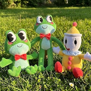 Könnte beinhalten: Zwei grüne Plüschfrösche mit großen schwarzen Augen, rosa Wangen und roten Fliegen stehen neben einer gelb-weißen Plüsch-Clownpuppe mit blauem Rüschenkragen, roten Knöpfen und roten Schuhen, alle zusammen auf einer grünen Wiese platziert.