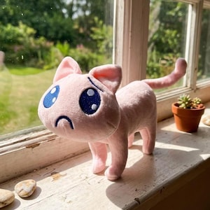 Pode incluir: Um brinquedo de pelúcia de gato rosa claro com grandes olhos bordados em azul escuro e uma pequena boca com expressão triste. A figura felina, macia e acolchoada, apoia-se em quatro patas com uma longa cauda curvada para cima.