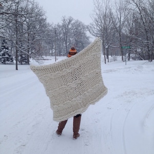 May include: A person walking in the snow, holding a large, cream-colored knitted blanket. The blanket features a cable knit design. The person is wearing brown boots and an orange and black hat. The scene is outdoors on a snowy day.