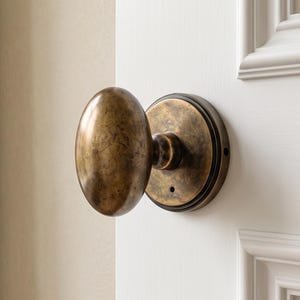 May include: An oval-shaped door knob with a distressed antique brass finish. The knob is mounted on a circular matching brass backplate and is attached to a white paneled door. The metal surface features visible dark oxidation and texture.