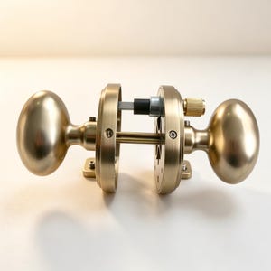 May include: A brushed brass door knob set featuring two oval-shaped handles attached to circular mounting plates. The mechanism includes a central square spindle, a black plastic component, and a small knurled brass adjustment screw on the side.