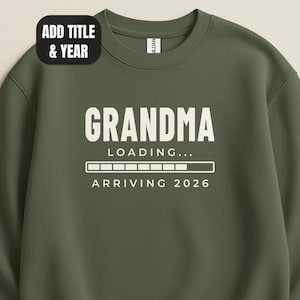 May include: An olive green crewneck sweatshirt featuring white text that reads GRANDMA LOADING... ARRIVING 2026 with a progress bar graphic. A black rectangular badge in the upper left corner contains the white text ADD TITLE & YEAR.