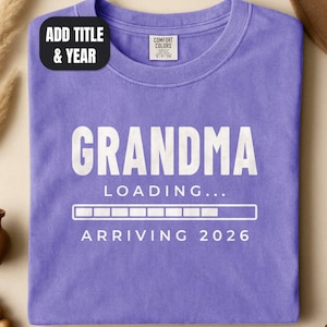 May include: A purple crew neck t-shirt featuring white text that reads GRANDMA LOADING... ARRIVING 2026 with a progress bar graphic. The shirt has a Comfort Colors brand label at the inner neckline and a black tag reading ADD TITLE & YEAR.