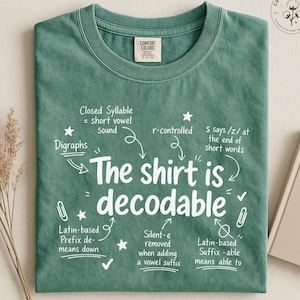 May include: A light green crew neck t-shirt featuring white text that reads The shirt is decodable, surrounded by educational linguistic notes including Closed Syllable, Digraphs, r-controlled, and Latin-based prefixes and suffixes with small star icons.
