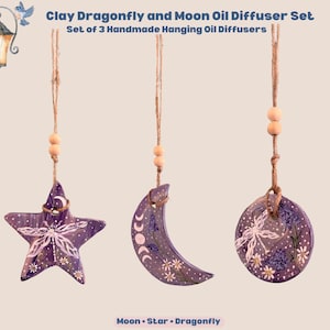 May include: A set of three purple clay hanging oil diffusers featuring a star, a crescent moon, and a circle shape. Each piece is decorated with hand-painted white dragonfly and floral motifs, suspended by twine with wooden beads. Text reads: Clay Dragonfly and Moon Oil Diffuser Set, Set of 3 Handmade Hanging Oil Diffusers, Moon, Star, Dragonfly.