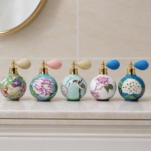 May include: Five round glass perfume bottles with gold-tone atomizers and fabric bulbs. The bottles feature colorful floral and bird designs, including a parrot on green, pink peonies on blue, a bird on light green, peonies on white, and white flowers on blue.