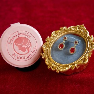 May include: A pair of drop earrings featuring oval red stones surrounded by small clear stones, displayed in an ornate gold-toned oval frame with a blue velvet interior, next to a round pink lid printed with the text Selena Jewelry & Bullion Inc.