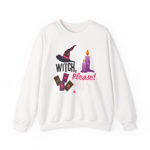 May include: A white long-sleeve sweatshirt featuring a graphic print of a black witch hat, two purple candles with orange flames, three tarot cards, a spider web, and a small spider. The text "WITCH, Please!" is printed in black and pink.