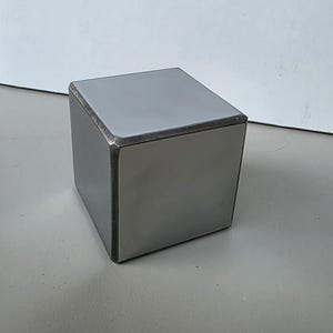 May include: A solid, metallic cube with a smooth, matte grey finish. The object features sharp, defined edges and a uniform, industrial appearance. The surfaces are flat and reflective, showing subtle variations in the grey tone across each side.