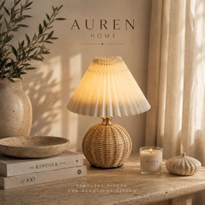 Puede incluir: Una lámpara de mesa con base redonda de ratán tejido y pantalla de tela blanca plisada. La lámpara emite una luz cálida. El texto AUREN HOME es visible sobre la lámpara, y el texto TIMELESS PIECES FOR BEAUTIFUL LIVING está debajo.