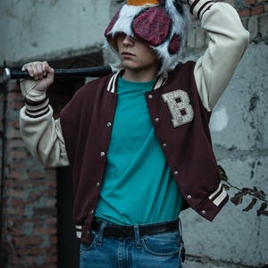 May include: A maroon and cream varsity jacket with a letter B patch, a teal t-shirt, blue jeans, and a black belt. A plush chicken head mask with white, black, and red details is worn on the head, and a black baseball bat is held across the shoulders.