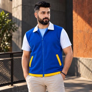 Könnte beinhalten: Eine blaue ärmellose Weste mit Reißverschluss, weißem Kragen und gelben Akzenten an den Taschen und am Saum, getragen über einem weißen kurzärmeligen Hemd und einer hellen Hose. Die Weste hat einen durchgehenden blauen Reißverschluss.