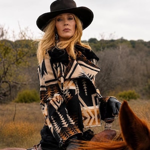 May include: A black felt wide-brimmed hat and a cropped jacket featuring a tan, black, and brown geometric pattern with a wide collar. The jacket is worn with black leather gloves, and the ensemble is paired with dark trousers.