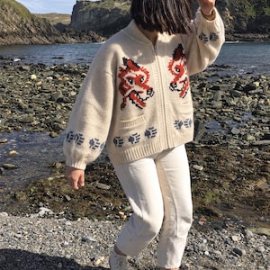 以下が含まれることがあります： 胸元に赤と黒のキツネの頭のモチーフ、袖と裾に青い足跡の模様があしらわれたクリーム色のニットジップアップカーディガン。白いクロップドパンツと、ガムラバーソールのオフホワイトのキャンバススニーカーを合わせています。