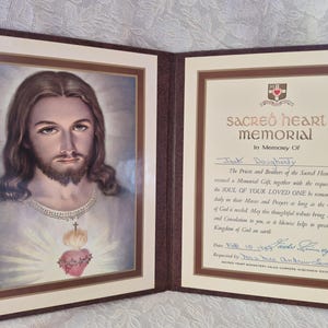 May include: A bi-fold memorial folder featuring a portrait of Jesus with a Sacred Heart on the left panel and a printed Sacred Heart Memorial certificate on the right panel. The certificate includes the text Sacred Heart Memorial in Memory Of.