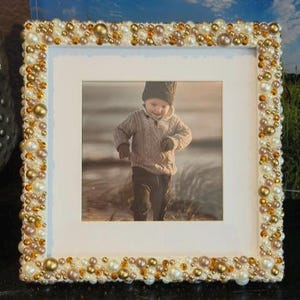 May include: A square picture frame featuring a white border densely decorated with an assortment of gold, bronze, and white pearls and beads of varying sizes. The frame contains a photograph of a child wearing a knit sweater and hat.