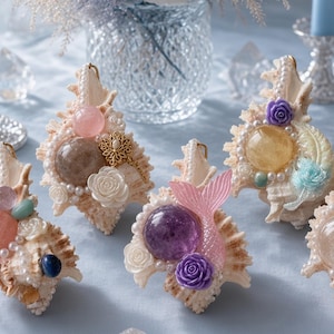Pode incluir: Uma coleção de conchas decorativas adornadas com vários elementos, incluindo pedras preciosas esféricas polidas, pequenas rosas de resina brancas e roxas, fios de pérolas falsas e uma estatueta de cauda de sereia rosa translúcida.