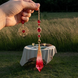 Puede incluir: Un adorno colgante con cadena dorada, cuentas rojas y una piedra roja ovalada. Una hebra termina en un dije de estrella dorada, mientras que la otra presenta un largo colgante puntiagudo de cristal translúcido rojo y blanco.