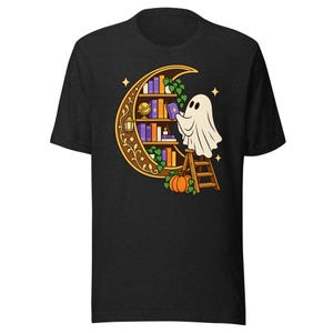 Peut inclure: Un t-shirt noir à manches courtes avec un motif représentant un fantôme blanc sur une échelle en bois, attrapant un livre sur une étagère en forme de croissant de lune. Le dessin inclut un globe, une lanterne, des vignes et une citrouille.
