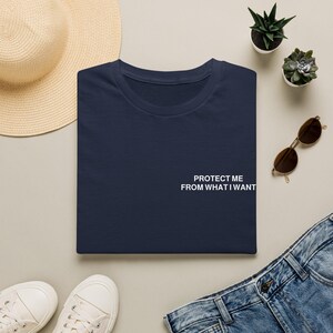 Puede incluir: Una camiseta azul marino con el texto blanco "PROTECT ME FROM WHAT I WANT" impreso en el lado derecho del pecho. La camiseta tiene cuello redondo y mangas cortas, presentada en una vista plana sobre un fondo neutro.