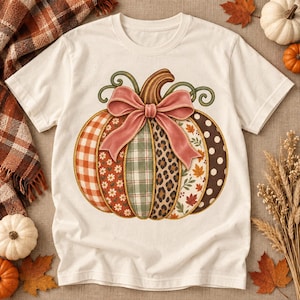 May include: A cream-colored t-shirt featuring a large central graphic of a pumpkin. The pumpkin is divided into vertical segments with various patterns including orange gingham, floral, green plaid, leopard print, and autumn leaves, topped with a pink bow.