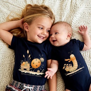 May include: A dark navy blue t-shirt featuring a ladybug pulling a wagon with pumpkins and the text "LITTLE HARVEST HELPER" and a dark navy blue baby bodysuit featuring a caterpillar with a scarf and the text "STILL IN MY COCOON".