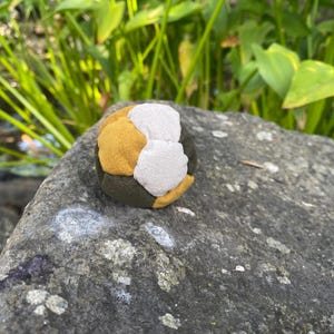 May include: A spherical juggling ball constructed from multiple fabric panels in shades of mustard yellow, off-white, and dark olive green. The ball features a soft, textured surface with visible stitching connecting the individual colored fabric segments.