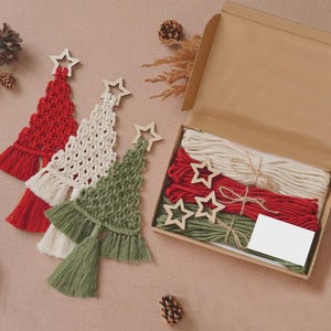 Puede incluir: Tres adornos de macramé con forma de árbol de Navidad en rojo, crema y verde salvia, cada uno con una estrella de madera. Junto a ellos, una caja de cartón abierta contiene cordones de algodón a juego y tres estrellas de madera.