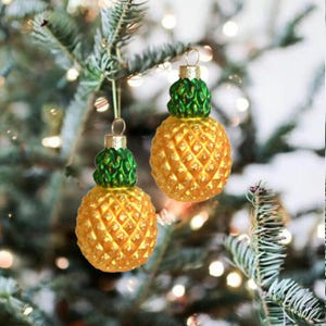 May include: Two pineapple-shaped ornaments featuring a textured golden yellow body and a vibrant green leafy top. Each ornament includes a small metallic cap at the crown with a loop for hanging, designed with a detailed cross-hatch pattern.