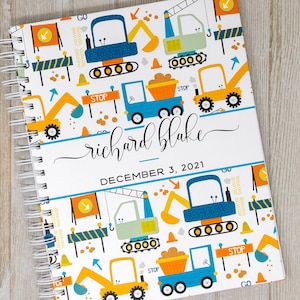 May include: A white spiral bound baby book with a construction theme. The cover features a repeating pattern of yellow, blue, and green construction vehicles, stop signs, and traffic cones. The book is personalized with the name "richard blake" and the date "DECEMBER 3, 2021".