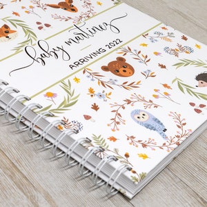 May include: A white spiral bound journal with a woodland animal theme. The cover features a variety of animals including foxes, owls, deer, and hedgehogs. The text on the cover reads "baby martinez arriving 2022".