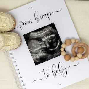 May include: A white journal with the text "from bump to baby" in black cursive script. The journal is open to a page with a black and white ultrasound image and a wooden baby teether with beads.