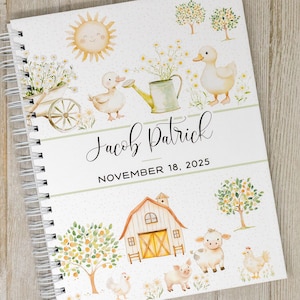 May include: A white spiral bound journal with a watercolor illustration of a farm scene. The cover features a yellow sun, a red barn, a duck, a pig, a cow, and a chicken. The text "Jacob Patrick" and "November 18, 2025" is printed on the cover.