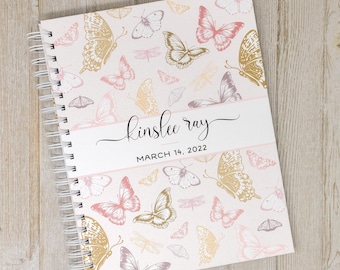 May include: A pink and white spiral bound journal with a butterfly pattern. The cover has the name "kinslee ray" and the date "March 14, 2022" printed on it.