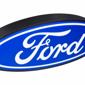 May include: An oval-shaped Ford sign featuring a blue background with a white border and the white Ford script logo in the center. The sign has a black outer edge and is presented at an angle to show its depth and three-dimensional construction.