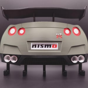 Könnte beinhalten: Ein olivgrüner Wand-Schlüsselhalter in Form eines Sportwagenhecks. Er verfügt über einen großen schwarzen Heckspoiler, vier runde rote Rückleuchten, ein GT-R-Emblem, ein weißes Nummernschild mit der Aufschrift nismo und vier schwarze Haken.