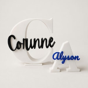 May include: A large white letter C with the black cursive name Corinne attached to the front, positioned next to a smaller white letter A with the blue cursive name Alyson attached to the front. Both letters stand upright on flat bases.