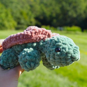 May include: A handmade crochet turtle plush toy featuring a soft, textured sage green body and a dusty rose pink shell. The turtle has a rounded head with a single visible black plastic eye and four small, rounded legs extending from the sides.
