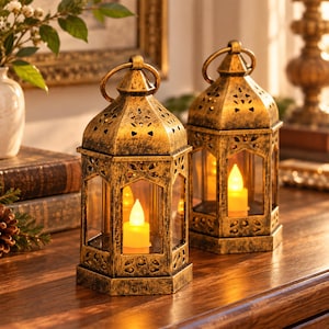 May include: Two hexagonal lanterns with a distressed gold finish and intricate cut-out patterns on the top panels. Each lantern features clear glass sides and a battery-operated LED candle inside that emits a warm, steady yellow glow.