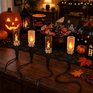 May include: A set of four electric candle lights with black bases and clear or frosted glass cylinders, each emitting a warm orange flame-like glow. The lights are connected by black cords and arranged on a dark wooden surface with Halloween decor.