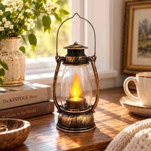 May include: A decorative lantern with a distressed black and gold finish featuring a clear glass globe and a metal handle. Inside the globe, a warm yellow flame-shaped light glows, mimicking a candle. The lantern has a sturdy base and top.