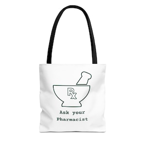May include: A white tote bag with black handles featuring a dark green line art illustration of a mortar and pestle with the Rx symbol inside. Below the graphic, the text reads Ask your Pharmacist in a dark green, typewriter-style font.
