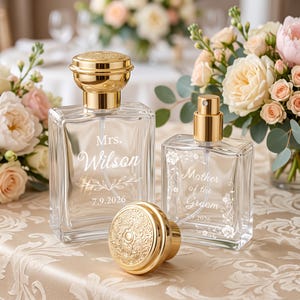 Può includere: Due flaconi di profumo in vetro trasparente con tappi dorati. Il flacone più grande è inciso con "Mrs. Wilson" e "7.9.2026". Il più piccolo è inciso con "Mother of the Groom" e "7.9.2026" insieme a dettagli di tralci floreali.