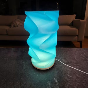 May include: A cylindrical table lamp with a bright cyan, spiraling, wave-like textured shade. The lamp features a circular base and a white power cord extending from the bottom, emitting a vibrant, glowing light from within the structure.