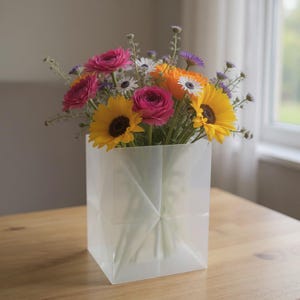 May include: A square, translucent white vase with a geometric folded design holds a vibrant bouquet of flowers. The arrangement features bright yellow sunflowers, deep pink ranunculus, orange blossoms, and small purple and white filler flowers.