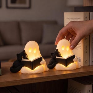 May include: Two small, glowing white ghost-shaped lamps, each featuring a black cat sitting beside it while holding an open black book. The ghosts have hollow, illuminated eyes and mouths, casting a warm light on the books and the surrounding shelf.