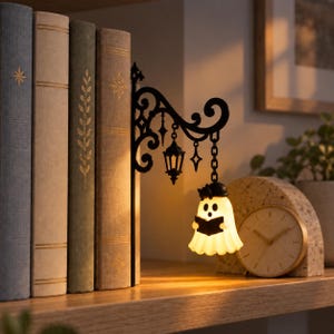 May include: A black metal bookend featuring an ornate scrollwork bracket with a hanging lantern and a small, illuminated white ghost figure wearing a black hat and holding an open book, positioned next to a row of four hardcover books.