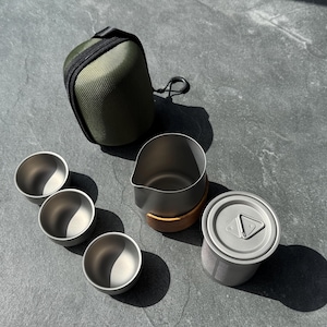 Könnte beinhalten: Ein Set aus drei kleinen Metallbechern, einem Metallkrug mit Holzboden, einem Metalldosenbehälter mit Deckel und einer olivgrünen Reißverschlusstasche. Die Gegenstände bestehen aus mattgrauem Metall und haben ein schlichtes, funktionales Design.
